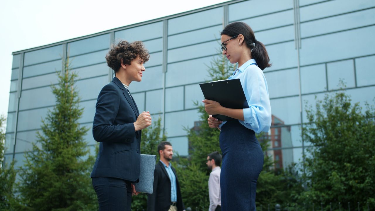 Two professional women discuss business outdoors, with modern building background.